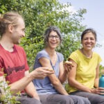 Drei Personen sitzen auf einer Holzbank in einer grünen, bewachsenen Umgebung und essen gemeinsam Snacks, umgeben von dichtem Gestrüpp und Natur.