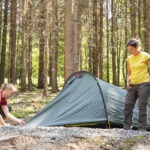 Zwei Personen richten im Naturpark Neckartal-Odenwald ein Trekkingzelt auf einer moosigen Waldlichtung zwischen hohen Fichten und Buchen auf.