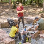 Drei Personen bereiten gemeinsam eine Mahlzeit auf einem Campingtisch aus Steinen im Wald des Naturparks Neckartal-Odenwald vor, Kochgeschirr und Rucksäcke liegen bereit.