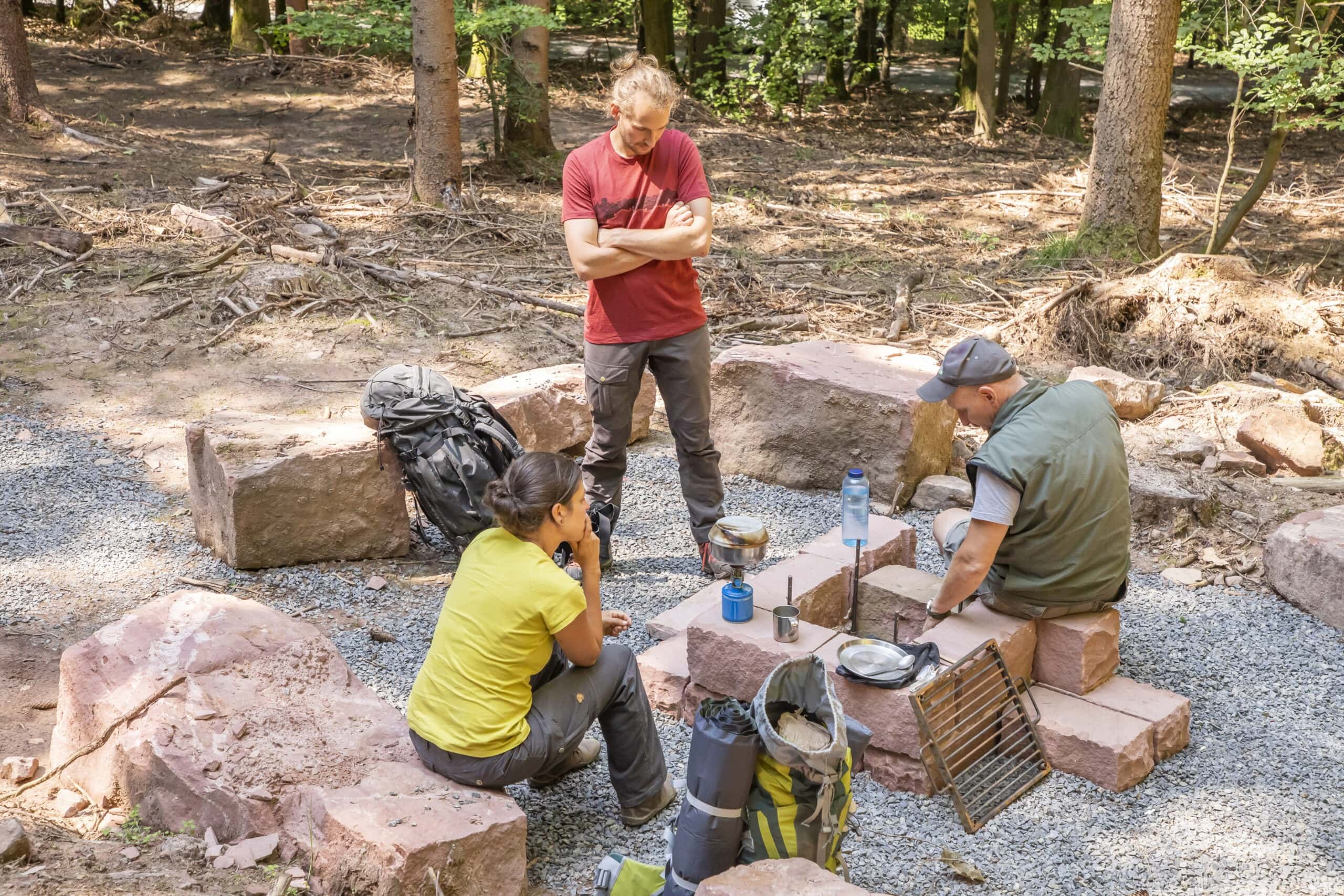 Drei Personen bereiten gemeinsam eine Mahlzeit auf einem Campingtisch aus Steinen im Wald des Naturparks Neckartal-Odenwald vor, Kochgeschirr und Rucksäcke liegen bereit.