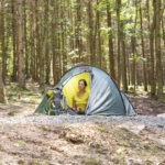 Person sitzt entspannt vor einem aufgeschlagenen Zelt im schattigen Wald des Naturparks Neckartal-Odenwald, umgeben von hohen Bäumen.