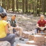 Drei Personen sitzen auf Steinen im Wald des Naturparks Neckartal-Odenwald, ein Zelt ist im Hintergrund aufgestellt, Kochgeschirr steht auf einem Campingtisch.