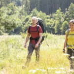 Zwei Wanderer mit Rucksäcken und Wanderstöcken durchqueren eine sonnige Wiese mit hohem Gras und wilden Blumen vor dichtem Wald im Naturpark Neckartal-Odenwald.