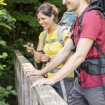 Zwei Wanderer mit Rucksäcken lehnen sich an ein Geländer im grünen Wald des Naturparks Neckartal-Odenwald, eine Person hält Trekkingstock.