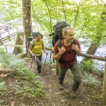 Zwei Wanderer mit Rucksäcken und Trekkingstöcken auf einem holzgesicherten Pfad im grünen Wald des Naturparks Neckartal-Odenwald, neben einem Bachlauf.