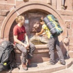 Zwei Wanderer am historischen Steinbrunnen im Naturpark Neckartal-Odenwald: Eine Person füllt eine Flasche, die andere sitzt mit Rucksack auf den Stufen.