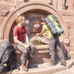 Zwei Wanderer am historischen Steinbrunnen im Naturpark Neckartal-Odenwald: Eine Person füllt eine Flasche, die andere sitzt mit Rucksack in einem steinernen Bogen.