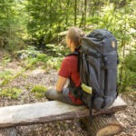 Person mit großem Trekkingrucksack sitzt auf einem Baumstumpf im Wald des Naturparks Neckartal-Odenwald und pausiert auf einem Waldweg.