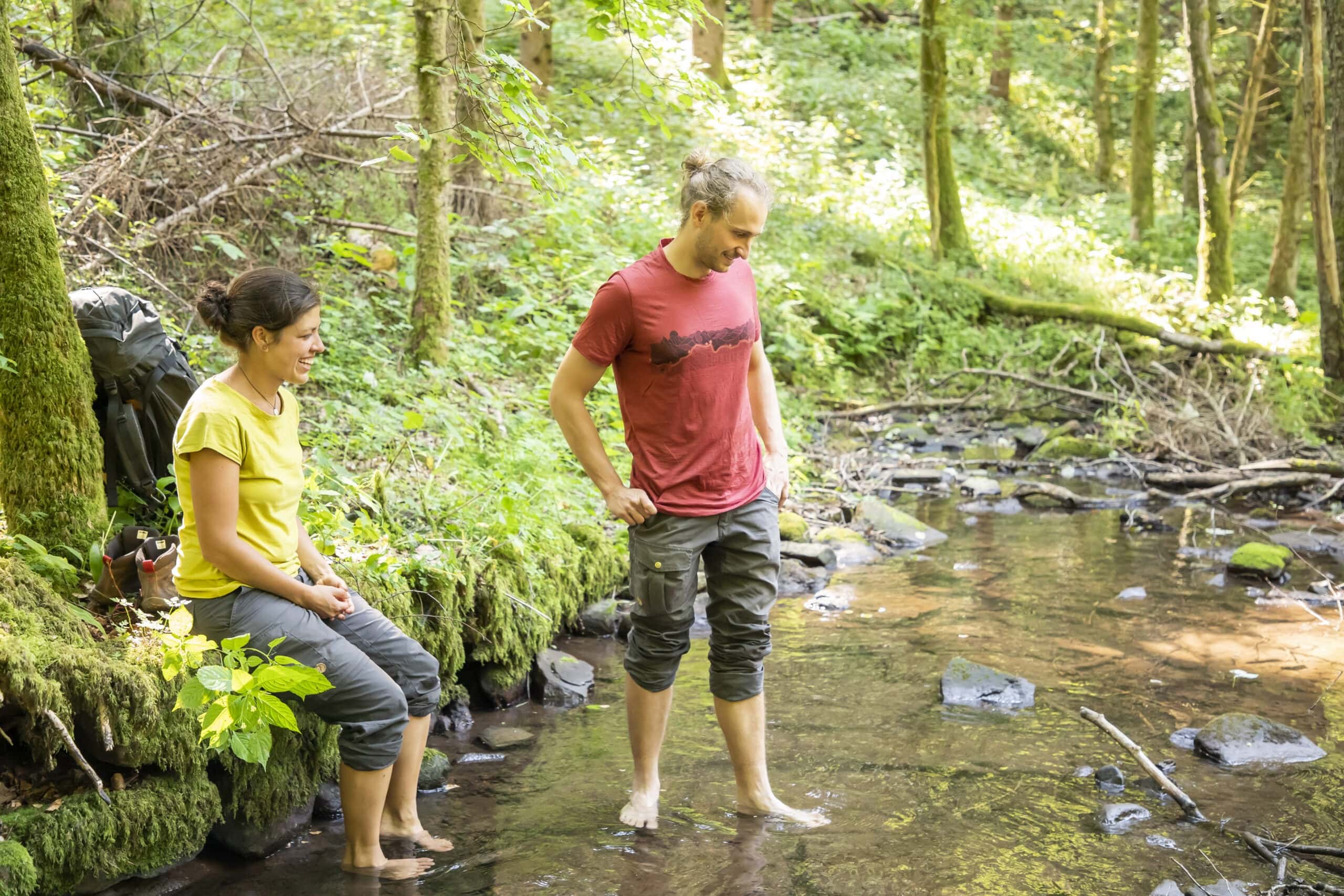 Zwei Personen am klaren Bach im Naturpark Neckartal-Odenwald: eine sitzt auf einem Baumstumpf, die andere steht barfuß im flachen Wasser, umgeben von moosbewachsenen Bäumen und Wald.