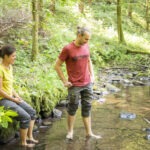 Zwei Personen am klaren Bach im Wald des Naturparks Neckartal-Odenwald: Eine sitzt auf einem Baumstumpf, die andere steht barfuß im flachen Wasser. Ein Rucksack liegt daneben.