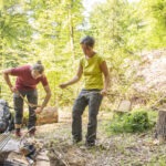 Zwei Personen sammeln im Wald des Naturparks Neckartal-Odenwald Äste für ein Lagerfeuer, während ein Rucksack und eine Grillvorrichtung im Hintergrund liegen.