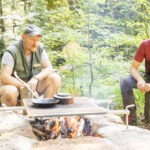 Zwei Personen sitzen am Lagerfeuer im Wald des Naturparks Neckartal-Odenwald, bereiten gemeinsam eine Mahlzeit auf einem Grillrost zu.