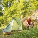 Person richtet grünes Zelt im schattigen Wald des Naturparks Neckartal-Odenwald auf natürlichem Untergrund ein, umgeben von dichtem Mischwald.