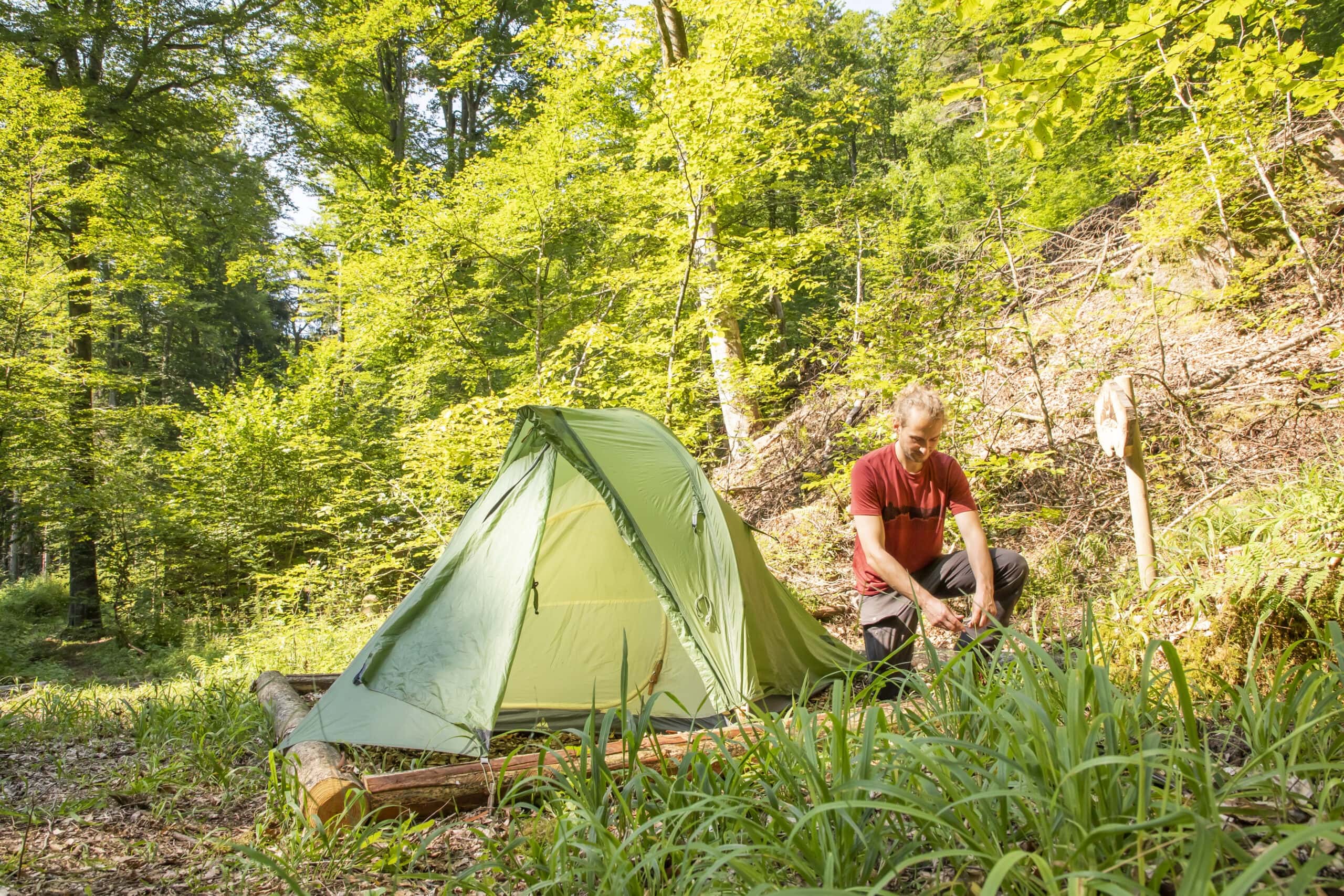 Person richtet grünes Zelt im schattigen Wald des Naturparks Neckartal-Odenwald auf natürlichem Untergrund ein, umgeben von dichtem Mischwald.