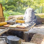 Feuerstelle im Naturpark Neckartal-Odenwald mit gegrilltem Fleisch, Maiskolben und Kartoffeln auf dem Rost, umgeben von Wald und natürlicher Umgebung.