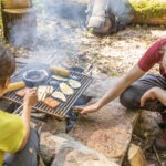 Zwei Personen grillen gemeinsam am Lagerfeuer im Naturpark Neckartal-Odenwald: gegrillte Maiskolben, Fleisch und Kartoffeln auf dem Rost, umgeben von Wald und natürlicher Umgebung.
