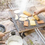 Grillen auf Campinggrill im Wald: Gegrillte Maiskolben, Fleischstücke und Brot, daneben Kochutensilien wie Pfanne und Topf auf Steinuntergrund.