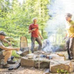 Drei Personen am Lagerfeuer im Wald: Eine kocht auf Campingkocher, zwei stehen und essen, umgeben von Steintischen und Baumstumpfsitzen.