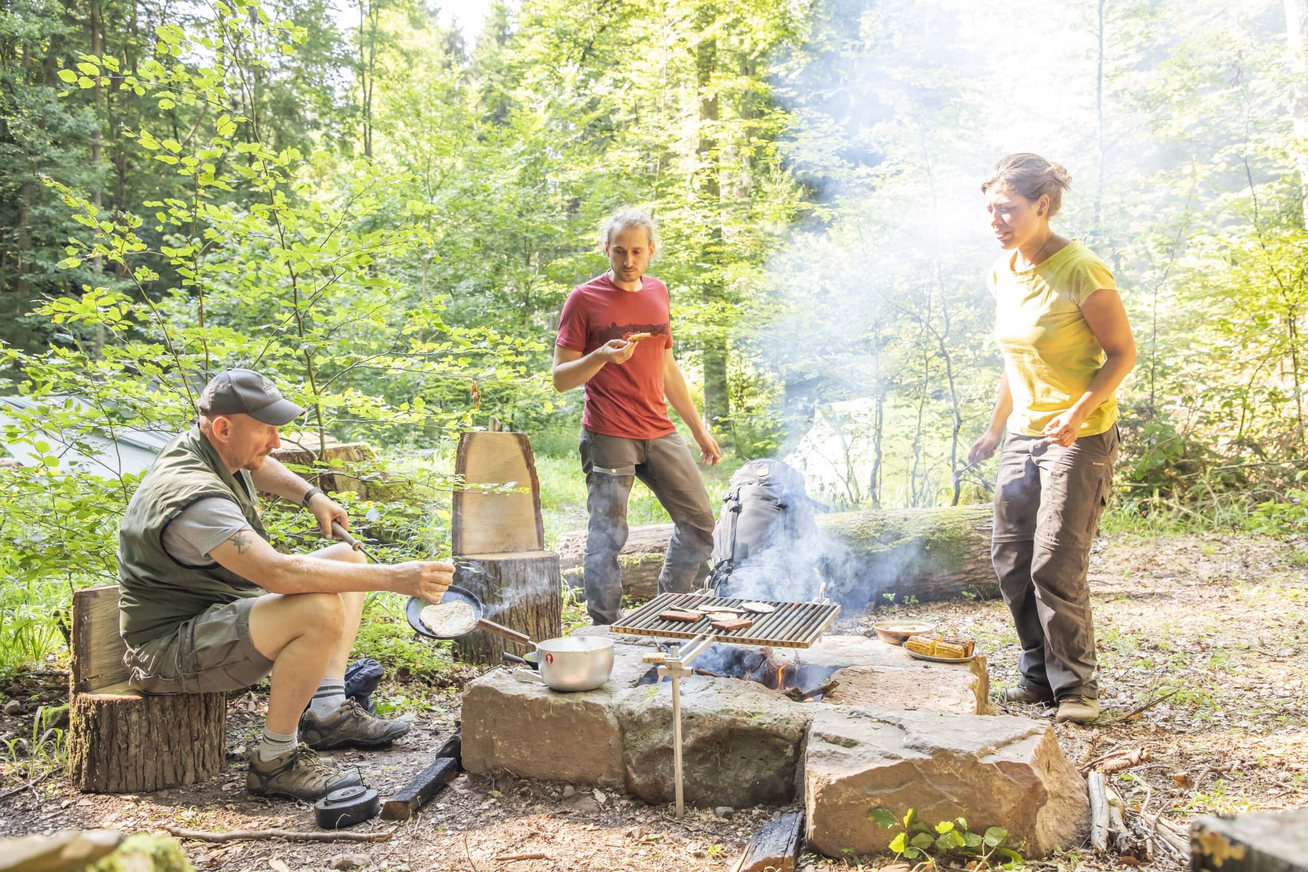 Drei Personen am Lagerfeuer im Wald: Eine kocht auf Campingkocher, zwei stehen und essen, umgeben von Steintischen und Baumstumpfsitzen.