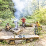Drei Personen am naturnahen Lagerfeuerplatz im Wald: Eine Person kocht, zwei sitzen auf Baumstämmen mit Rucksäcken und Ausrüstung.