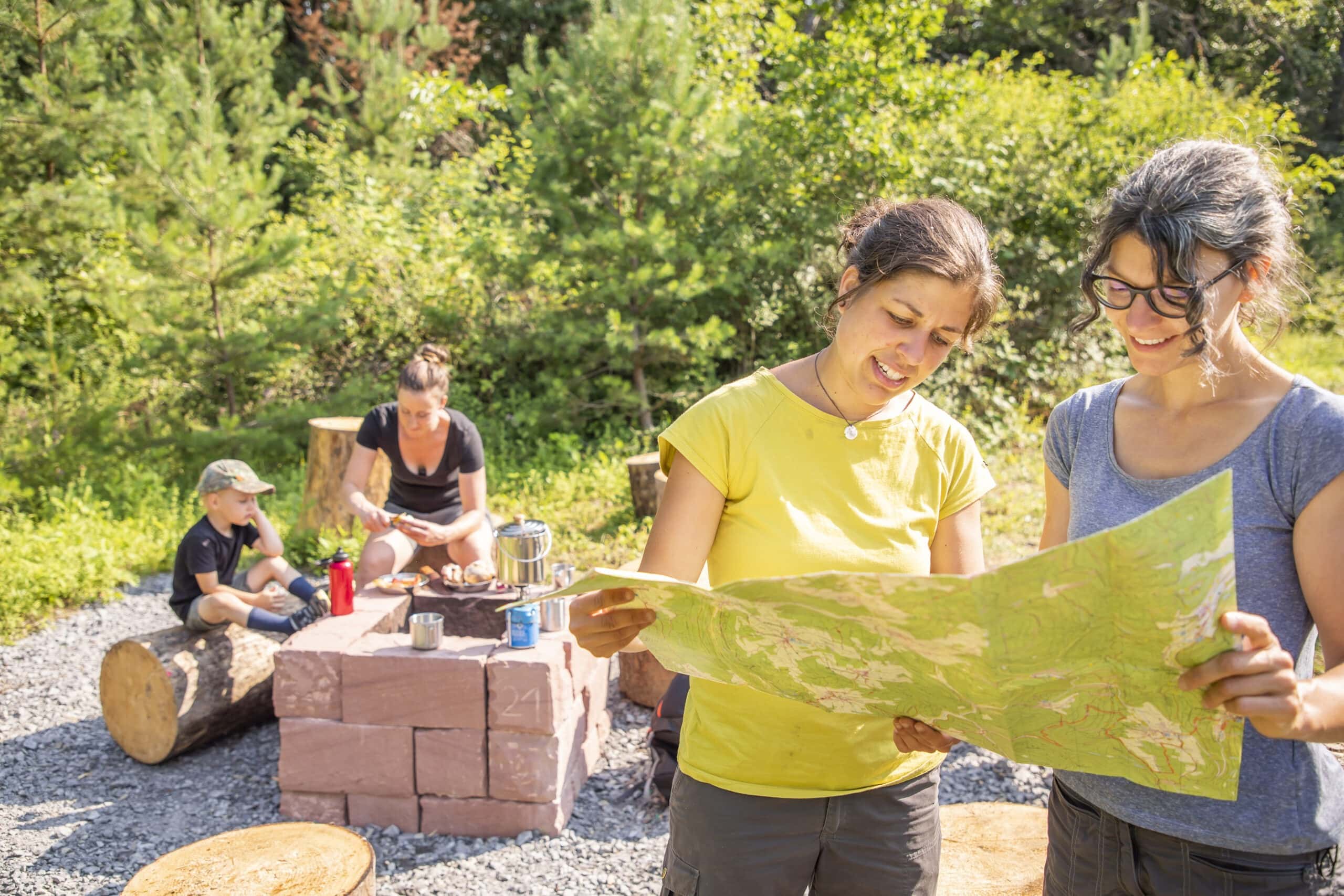 Zwei Personen studieren eine Wanderkarte am Trekking-Camp im Naturpark Neckartal-Odenwald, während andere am Tisch mit Kochutensilien sitzen.
