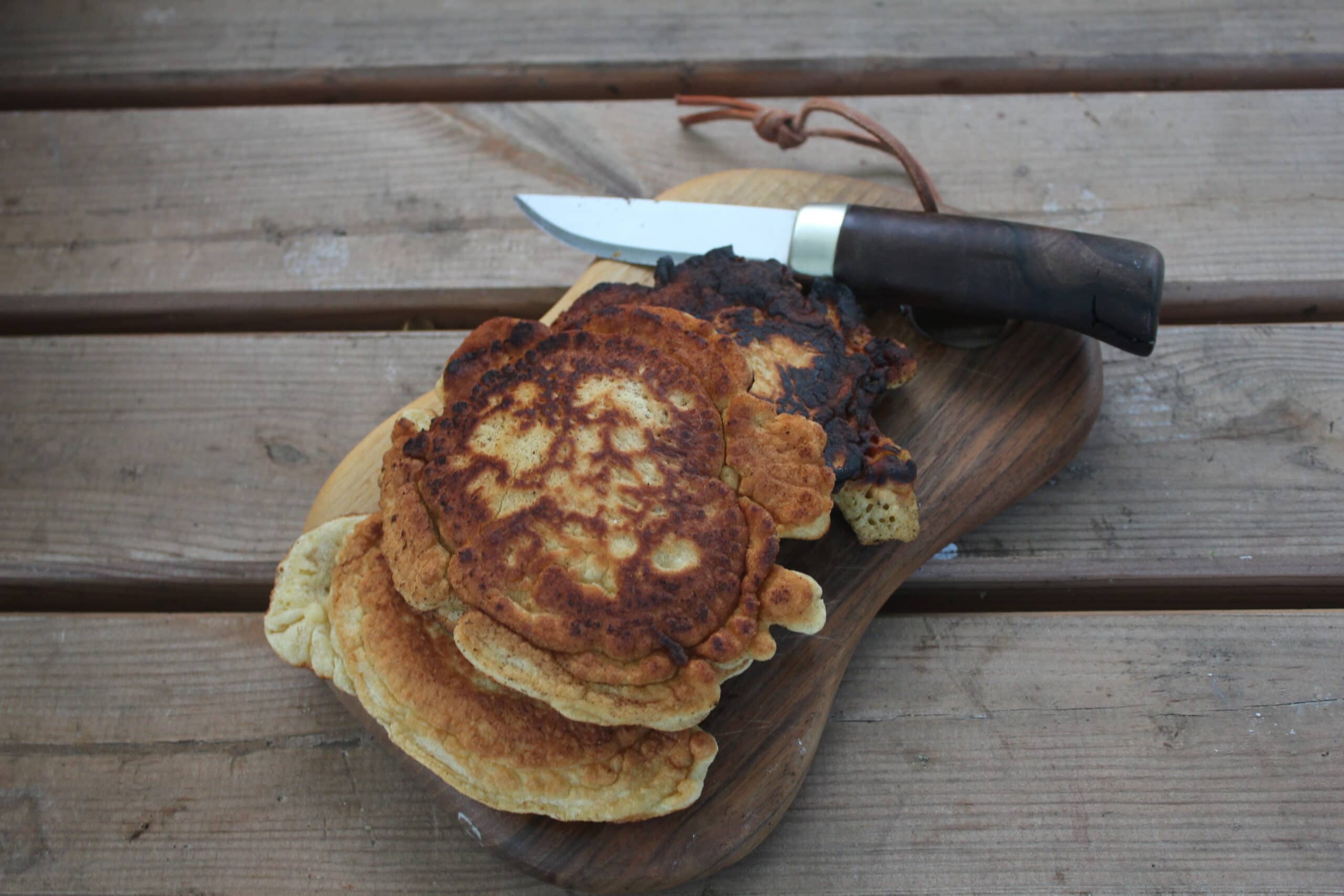 Gebratene Pfannkuchen auf einem Holzbrett mit Messer, platziert auf einer rustikalen Holzbank, typisch für einfache Mahlzeiten im Freien.