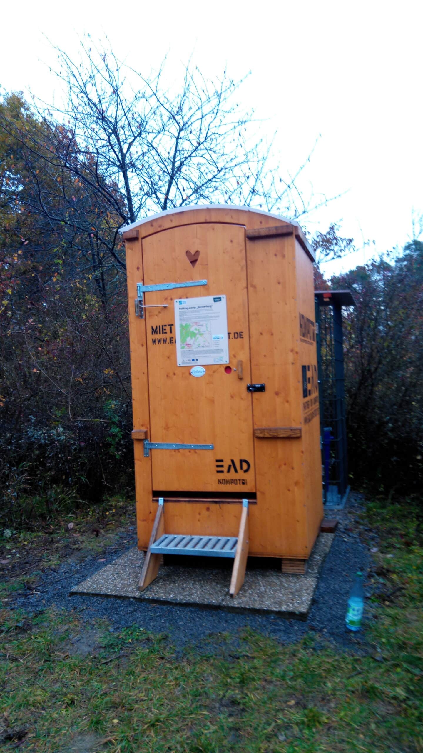 Holztoilettenhäuschen mit Treppe und Infoschild im Wald, umgeben von Sträuchern und Bäumen, typisch für naturnahe Wanderwege im Naturpark.