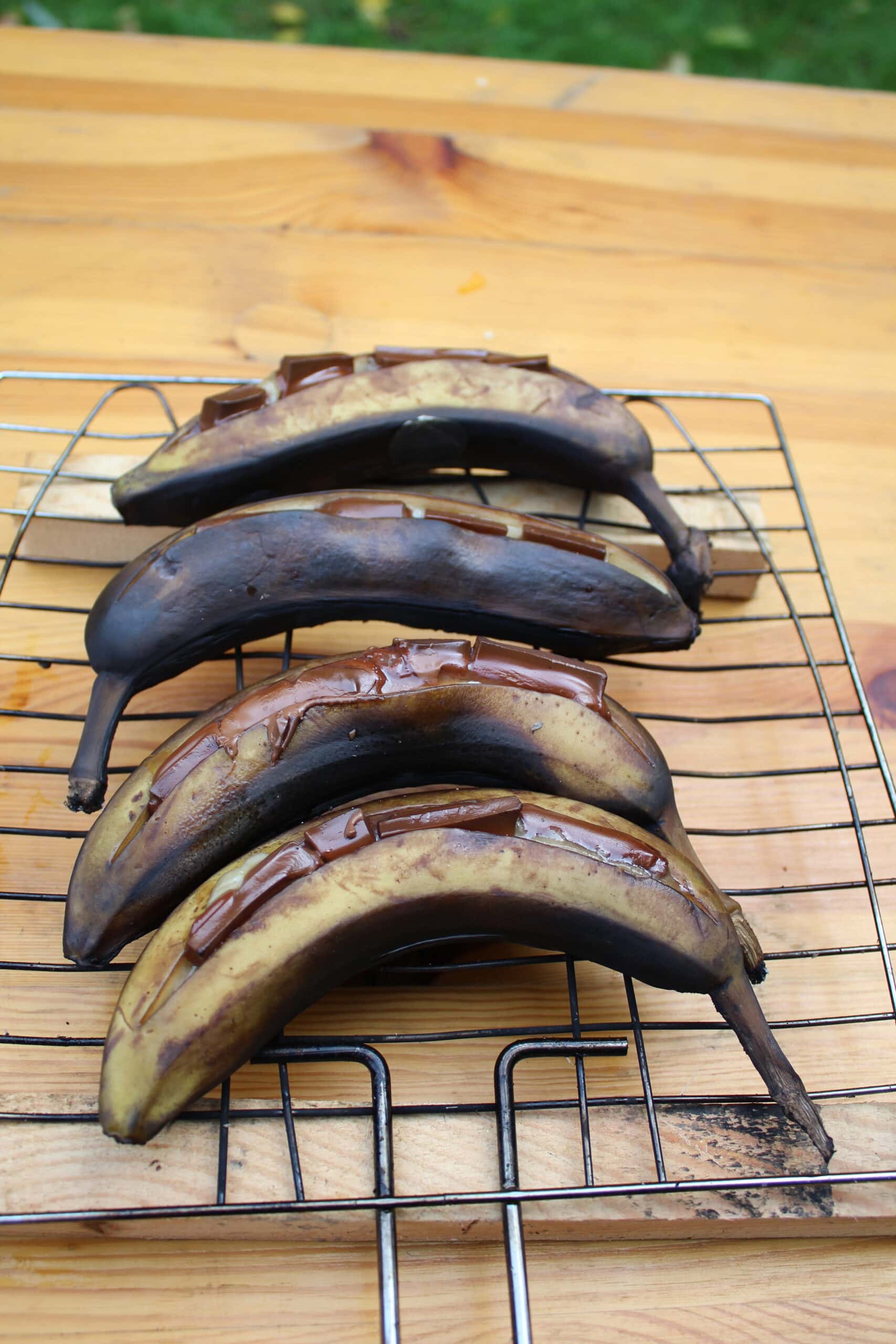 Vier gegrillte Bananen auf einem Drahtrost, leicht verkohlt, auf einer hellen Holzoberfläche.