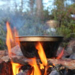 Feuerstelle im Naturpark Neckartal-Odenwald mit Metalltopf über Flammen, umgeben von Waldboden und Grünpflanzen im Hintergrund.