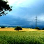 Weite Landschaft mit grünem Getreidefeld, einzelnen Bäumen und zwei Hochspannungsmasten unter bedrohlich wirkender Gewitterwolke am Horizont.