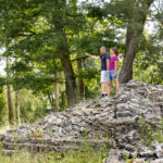 Zwei Personen stehen auf einer alten römischen Steinmauer im bewaldeten Odenwald, umgeben von dichtem Grün und Bäumen. Historische Limes-Reste im Naturpark Neckartal-Odenwald.