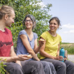 Drei Personen sitzen lächelnd auf einer Holzbank in einer grünen Odenwälder Landschaft, essen Snacks und genießen die Natur mit Blick auf Wiesen und Wald.