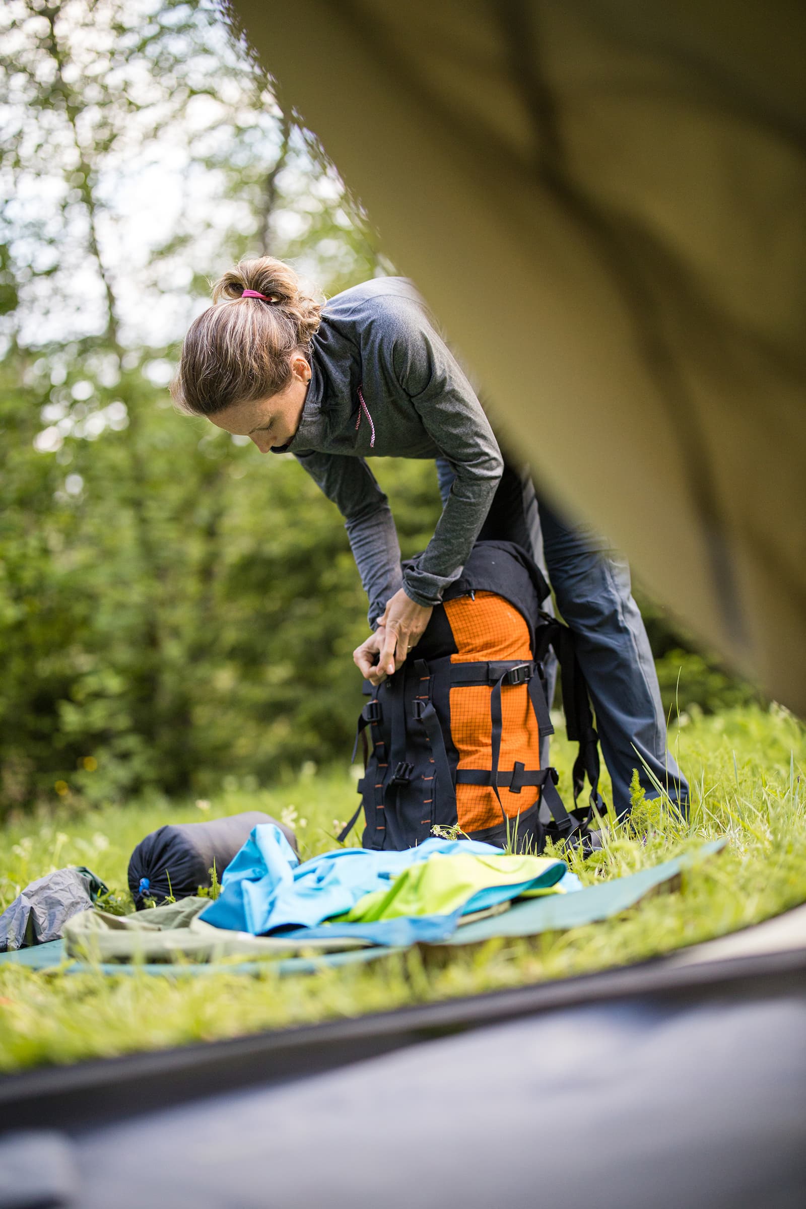Person packt im Wald Ausrüstung aus: Rucksack mit orangefarbenen Taschen, Isomatte und Schlafsack auf grüner Wiese.